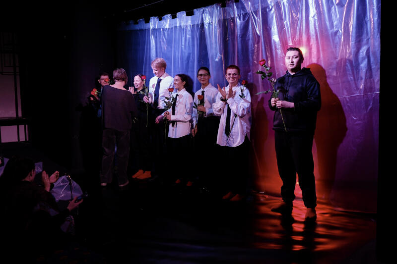 На слабо освещенной сцене с драпированным пластиковым фоном группа людей держит красные розы, в то время как аудитория аплодирует.