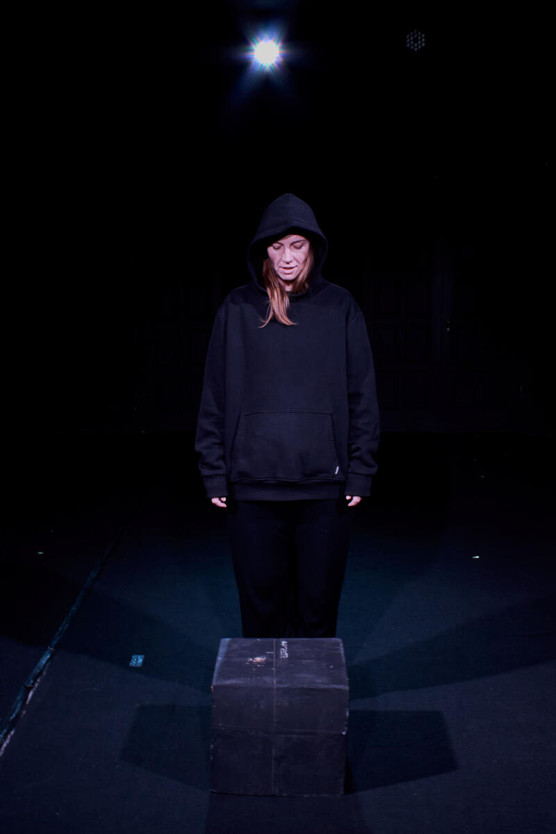 Женщина в черной толстовке с капюшоном стоит над коробкой под лучом света.