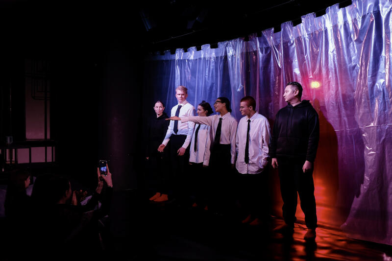 Группа актеров в белых рубашках и черных галстуках стоит на сцене перед полупрозрачным пластиковым фоном, освещенным цветными огнями, а зритель фотографирует происходящее на смартфон.