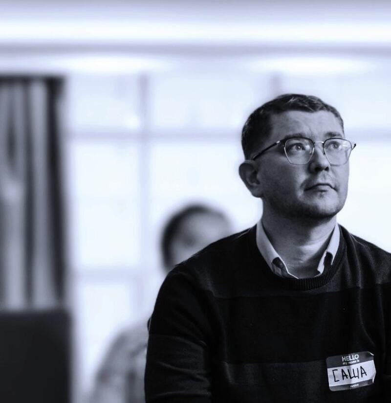 A man with glasses and a name tag that reads "ГАША" is wearing a striped sweater and looking up and to the right.