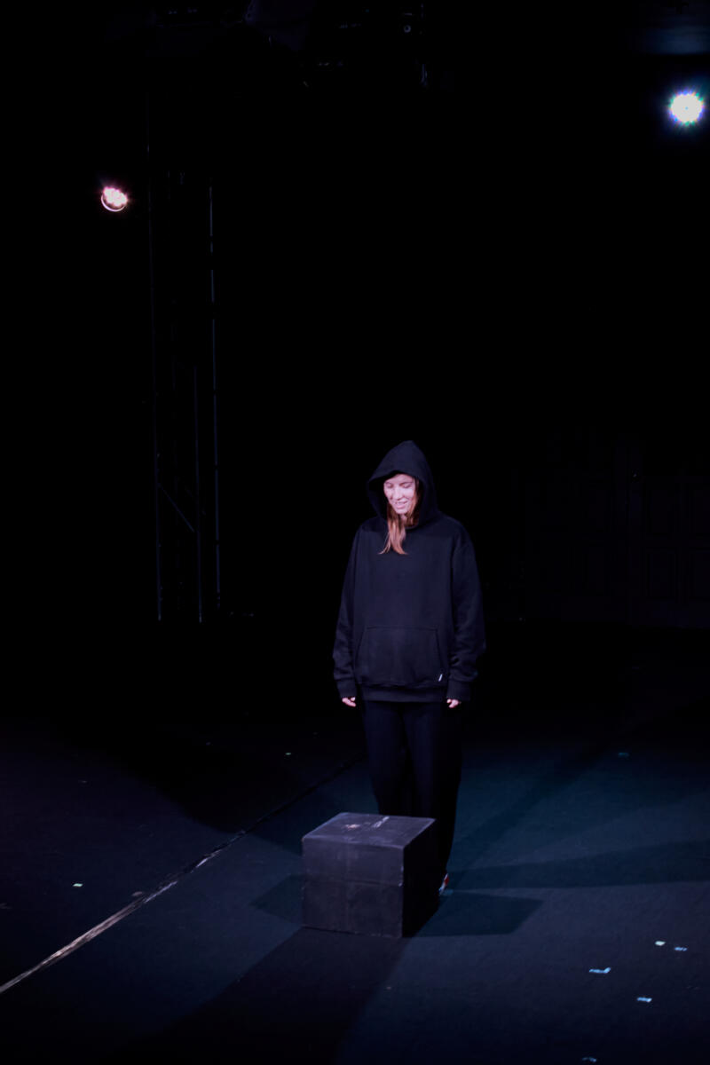 На фотографии изображена женщина в черном худи и черных брюках, стоящая на сцене рядом с черным кубом.