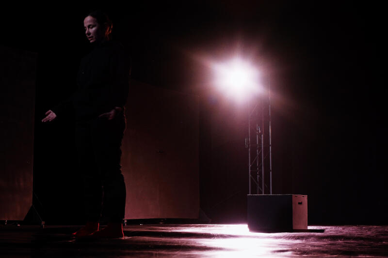 Женщина в темной одежде стоит на сцене, освещенной ярким прожектором, рядом с металлической вышкой и темным ящиком.