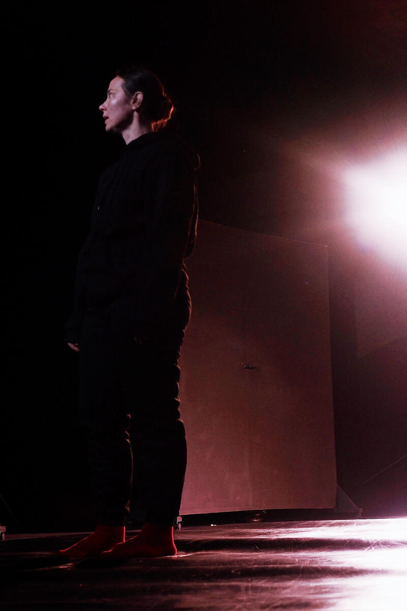 Женщина в темной толстовке и брюках стоит босиком на сцене, освещенной розовым светом.