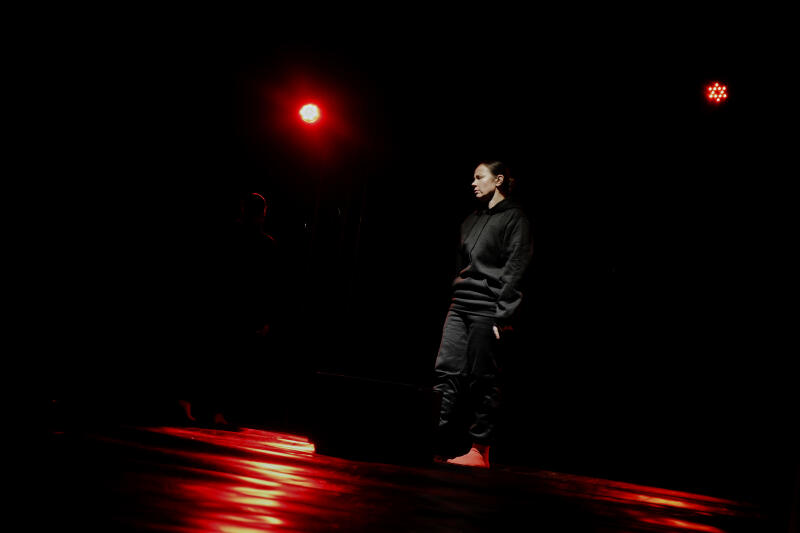 В слабо освещенной сцене стоит женщина, одетая в темную толстовку и брюки, освещенная красными сценическими огнями, которые отражаются от пола.