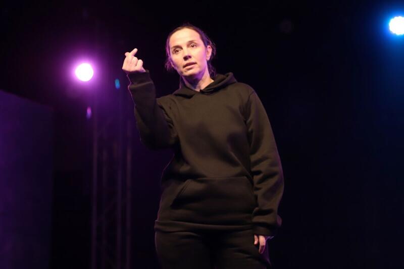 Женщина в черном худи стоит на сцене, делая жест «сердце пальцами», освещенная фиолетовым и синим сценическим светом.