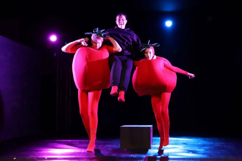 На сцене три исполнителя: двое одеты в красные костюмы помидоров с зелеными стеблями, а третий прыгает над ними в черной толстовке и штанах.