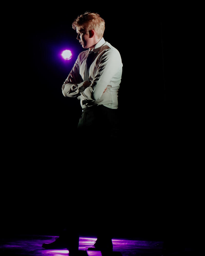 Мужчина с рыжеватыми волосами стоит, скрестив руки, освещенный фиолетовым прожектором в темной обстановке. На нем светлая рубашка, темные брюки и черные туфли.