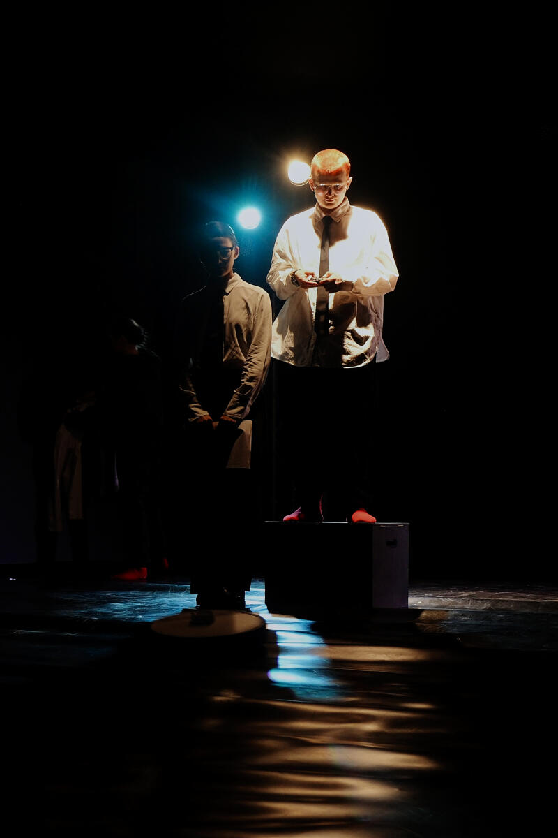 В полумраке человек с рыжеватыми волосами стоит на ящике, освещенный ярким светом, в то время как другой человек в очках стоит в тени с листом бумаги в руке.