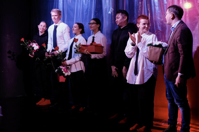 Группа людей стоит на сцене, некоторые держат цветы и подарки, на фоне драпированная ткань.