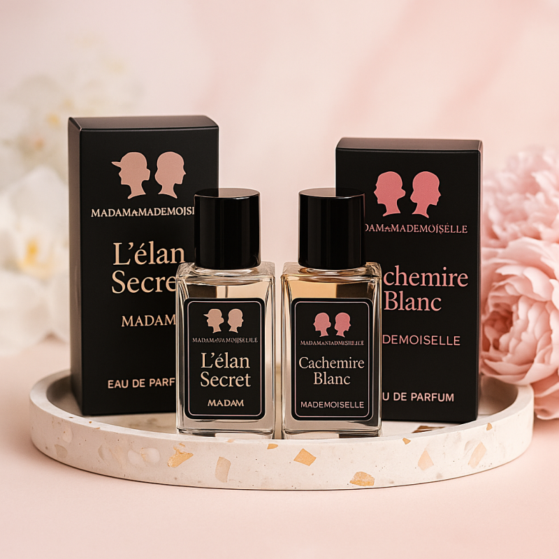 Two perfume bottles and their boxes, labeled "L'élan Secret MADAM" and "Cachemire Blanc MADEMOISELLE", are arranged on a speckled round tray, with pink flowers in the background.