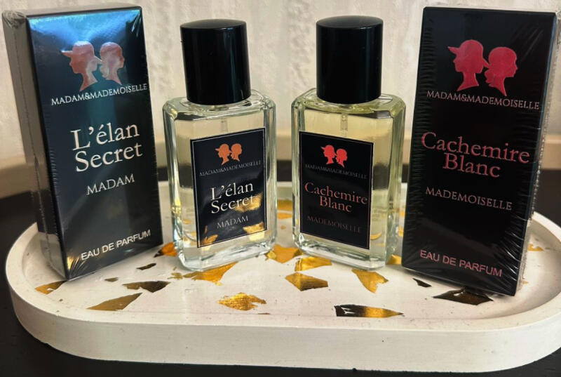 The image shows two perfume bottles and their boxes on a decorative tray with gold accents. The perfumes are labeled "L'élan Secret" and "Cachemire Blanc" by Madam&Mademoiselle.