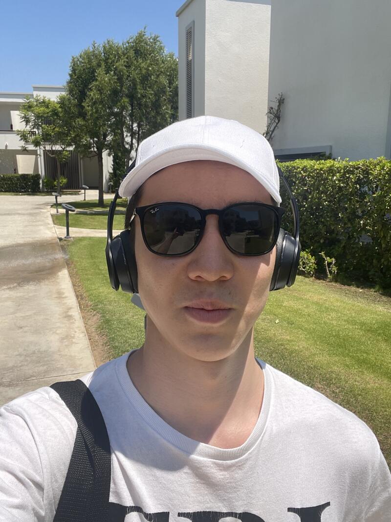 A person wears sunglasses, a white cap, and headphones, standing on a sidewalk with green grass and trees in the background.