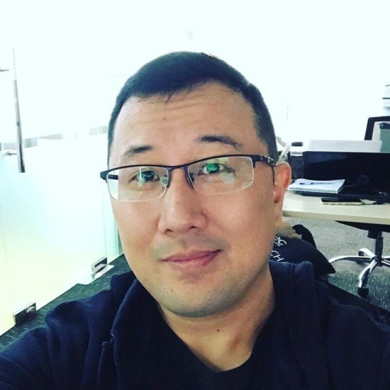 A close-up shot shows a man with short black hair and glasses, wearing a dark-colored hooded sweatshirt, in what appears to be an office setting.