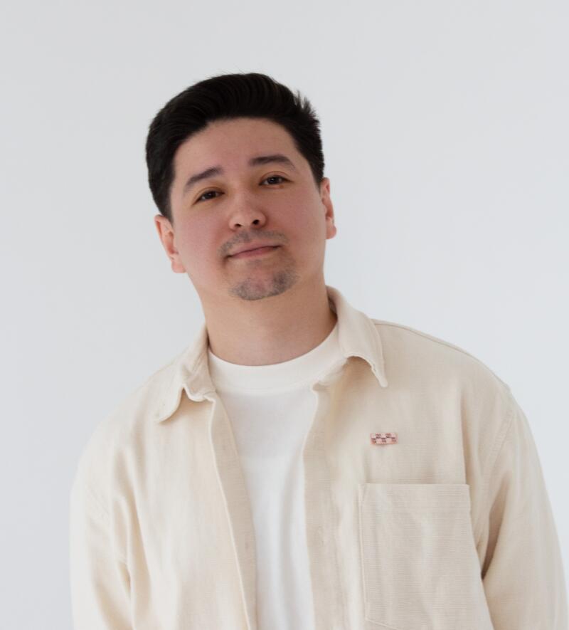 A man with dark hair and a goatee is wearing a white t-shirt and a tan button-up shirt with a small, checkered pin on the pocket.