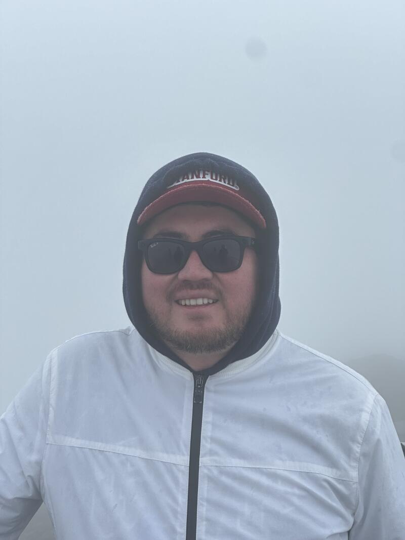 A person with sunglasses, a Stanford baseball cap, and a white jacket with a hood is smiling in a foggy environment.