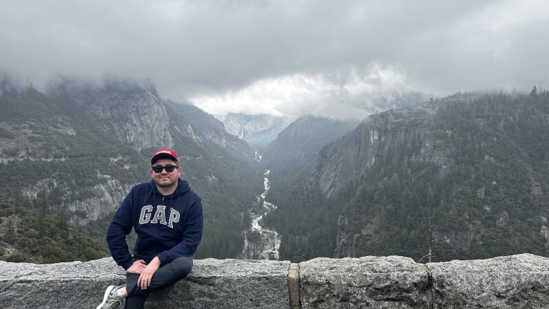 A man wearing a GAP hoodie, a red baseball cap, and sunglasses is sitting on a stone wall, with a mountain range in the background under a cloudy sky.