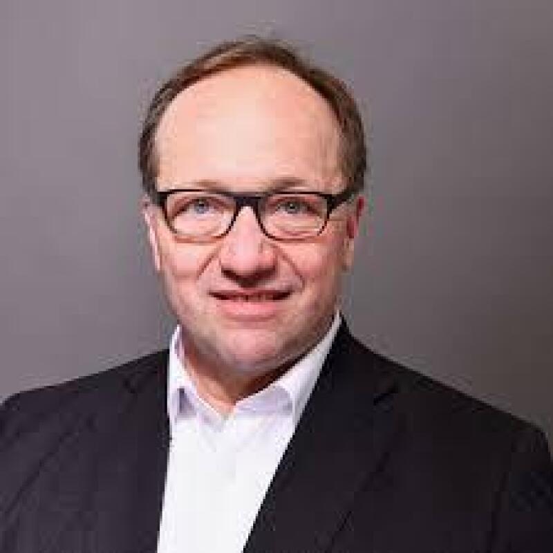 A man with glasses and a suit smiles at the camera against a gray background.