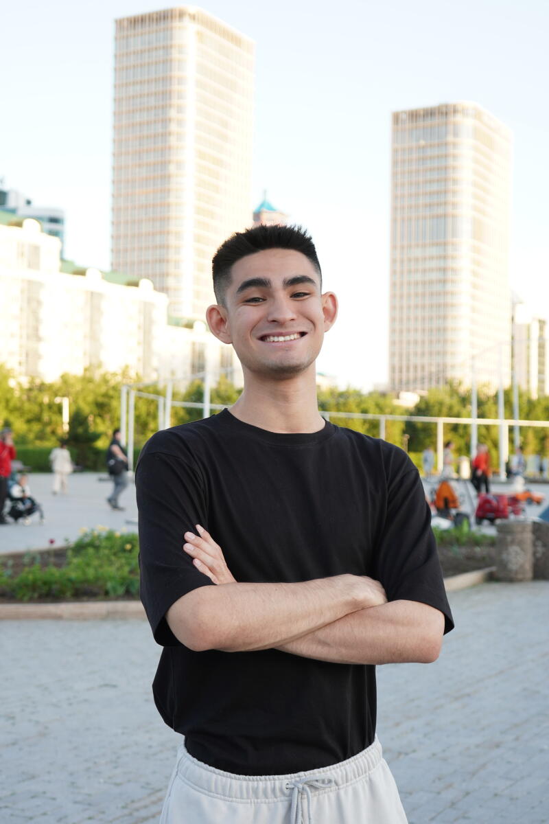A smiling man stands with crossed arms, wearing a black t-shirt and gray sweatpants, with tall buildings in the background.