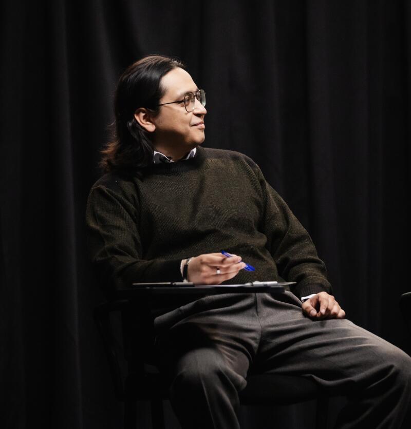 A man with long hair and glasses sits with a clipboard and pen, wearing a sweater and grey pants, against a dark curtain backdrop.