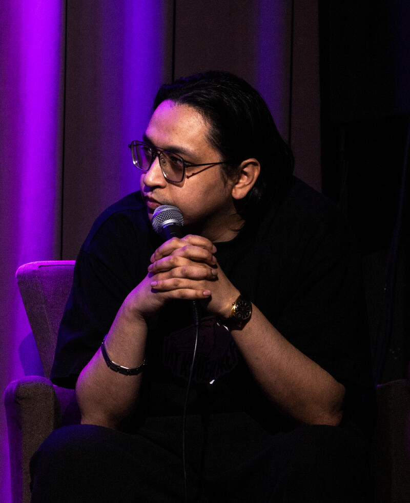 A man with glasses and long hair sits in a chair, holding a microphone with both hands. He is wearing a black T-shirt, a gold watch, and a bracelet.
