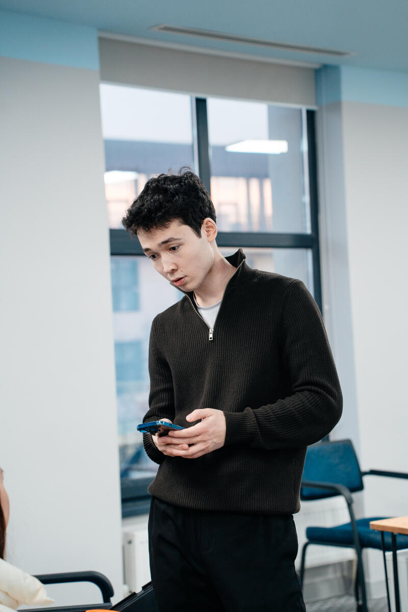 A young man with dark hair is looking at a blue smartphone in his hands, wearing a dark sweater and black pants, standing in a room with a large window.