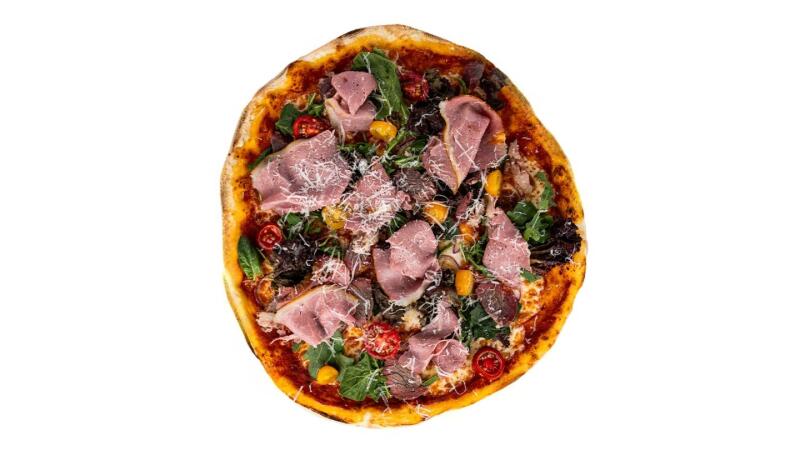 На фотографии сверху изображена пицца с золотистой корочкой, покрытая ветчиной, шпинатом, помидорами, желтым перцем и тертым сыром.