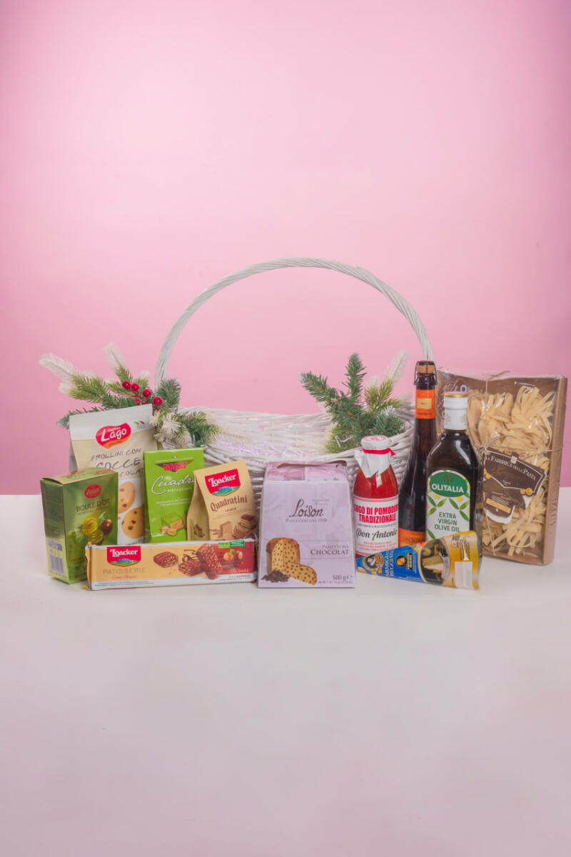 Подарочная корзина, наполненная различными продуктами питания, включая печенье, пасту, оливковое масло и томатный соус, украшенная искусственными сосновыми ветками, на розовом фоне.