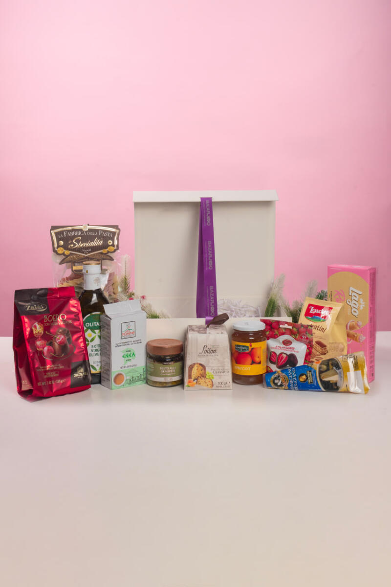 Подарочная коробка с различными продуктами питания представлена на розовом фоне, включая шоколад, оливковое масло, пасту, джем и вафли, коробка украшена фиолетовой лентой с надписью «BAZARJO».