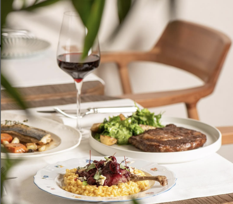 The photo shows a table setting with a glass of red wine, and three plates with different dishes: one with fish and vegetables, another with steak and salad, and a third with risotto and meat topped with cherries.