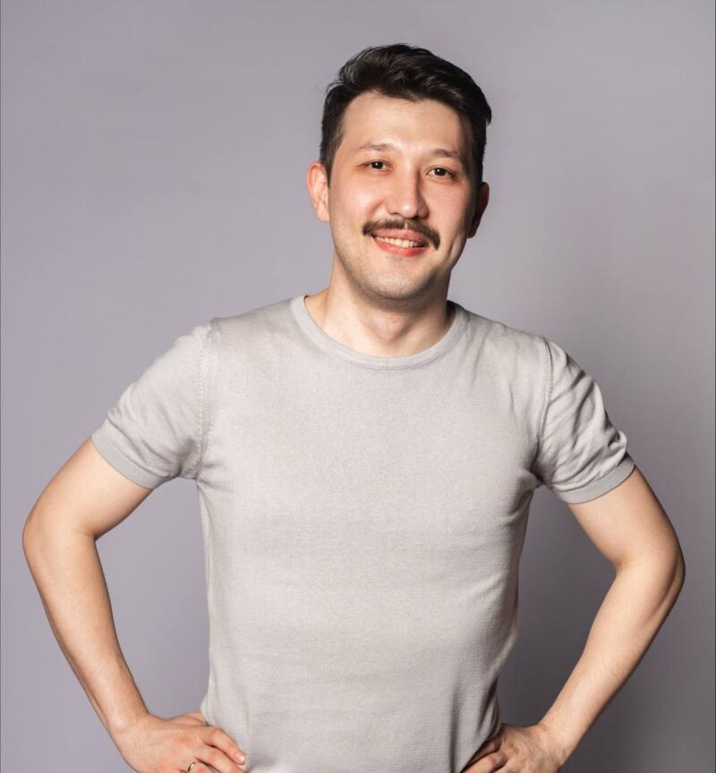 A man with a mustache is smiling and has his hands on his hips, wearing a gray t-shirt against a gray background.