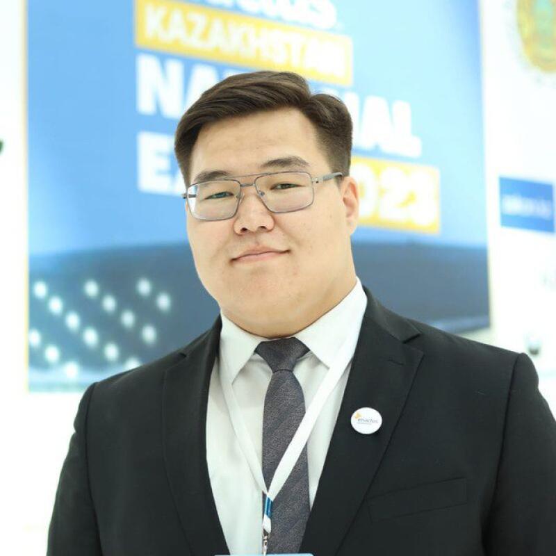 A man in a suit and tie stands in front of a poster that reads "KAZAKHSTAN NATIONAL 2023".