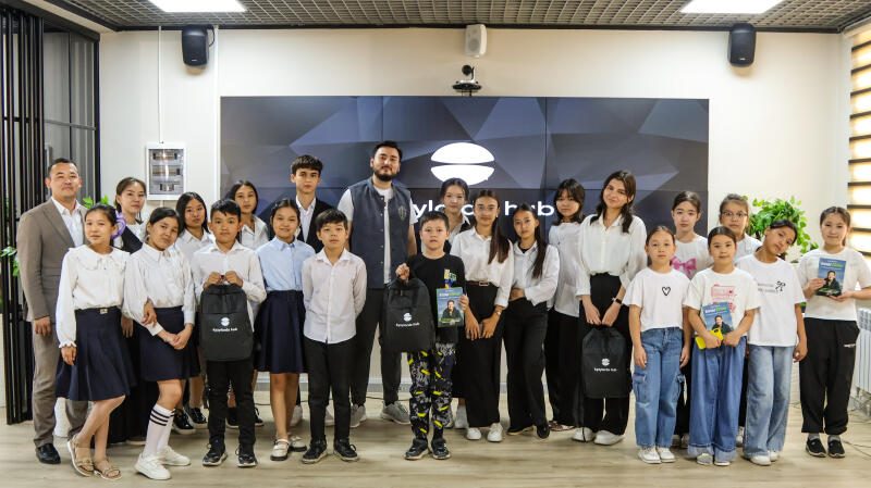 A group of approximately 20 children and two adults pose indoors, some holding books and backpacks with the "Myrzakent Hub" logo. The backdrop features a screen with the same logo and the words "Myrzakent Hub".