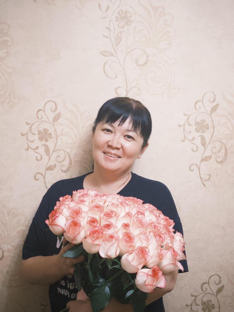 A smiling woman with short dark hair holds a large bouquet of light pink and white roses against a beige wall with a floral pattern.