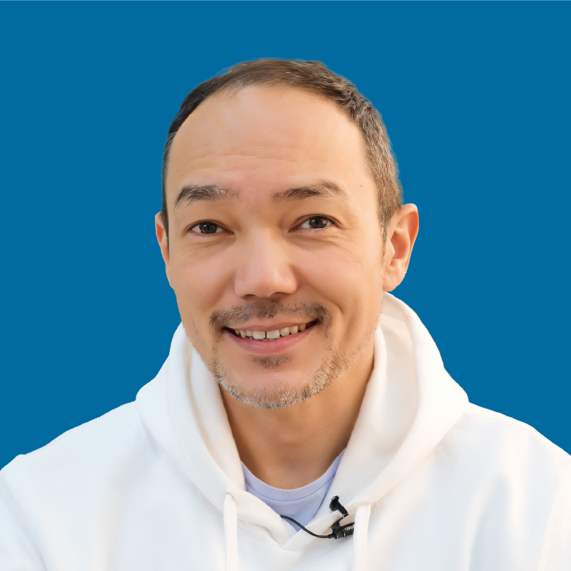 A man with short hair and a light beard is smiling and wearing a white hoodie against a blue background; a microphone is clipped to his hoodie.