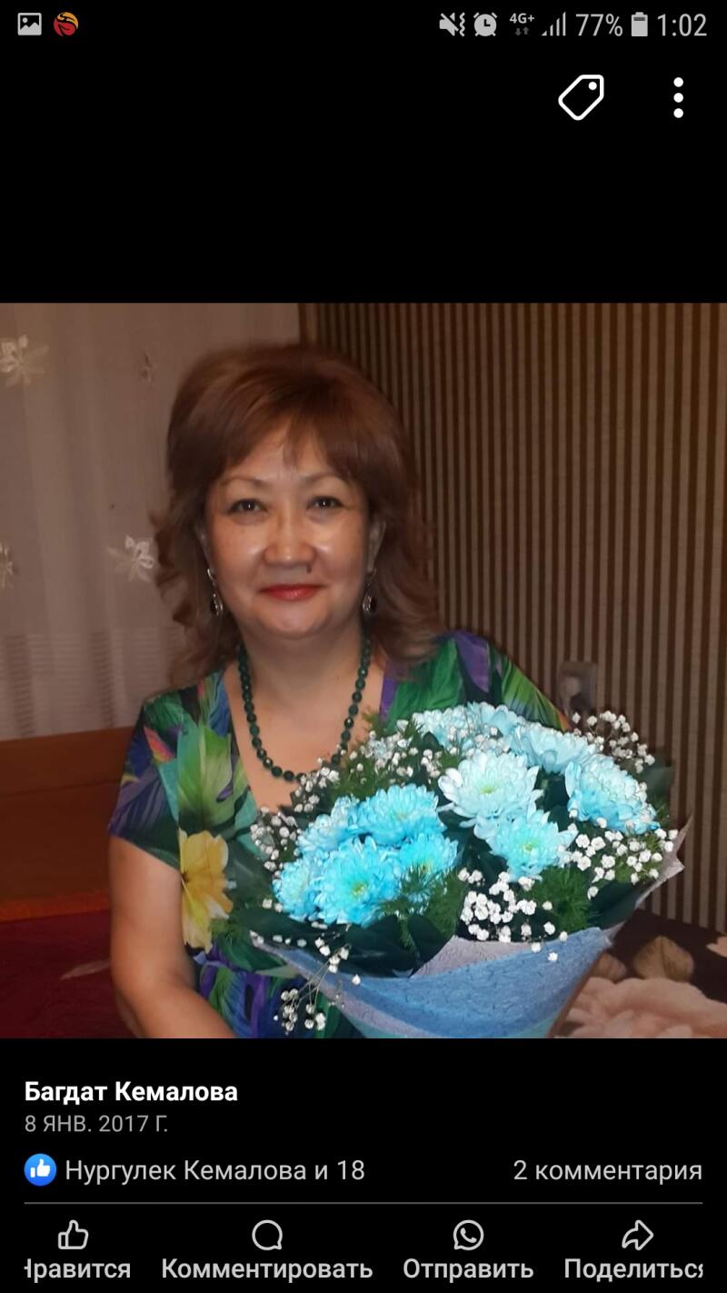 A woman with short brown hair is holding a bouquet of blue flowers and baby's breath, wearing a colorful top and green beaded necklace.