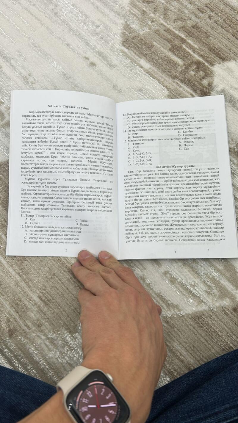 Человек держит открытой книгу с текстом на казахском языке, на левом запястье надеты смарт-часы, на страницах книги исторический контент и вопросы.