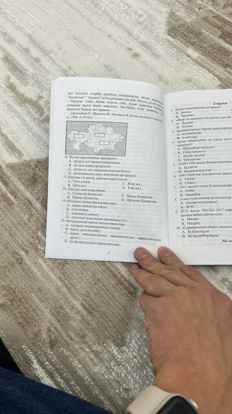 Видна открытая книга с текстом на казахском языке, содержащая параграфы и вопросы с вариантами ответов.