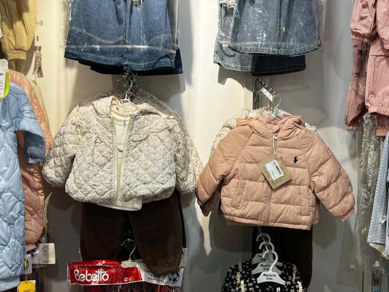 The image shows a display of children's clothing, including quilted jackets and denim skirts, hanging on racks.