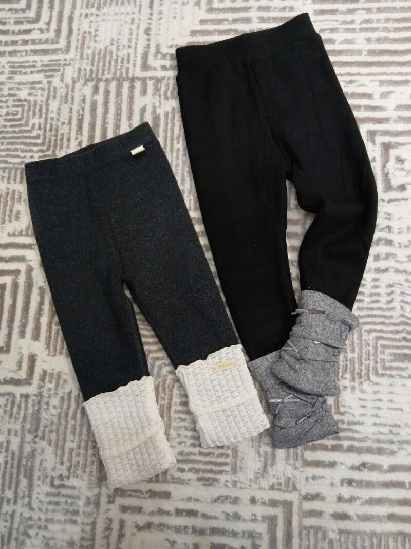 Two pairs of children's leggings, one dark gray with white cuffs and the other black with gray ruffled cuffs, are lying on a patterned surface.