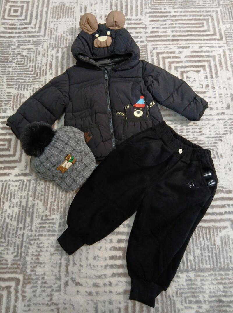 A children's set is displayed on a patterned surface, consisting of a black puffer jacket with bear ears on the hood, a gray plaid hat with a pompom and a bear patch, and black pants.