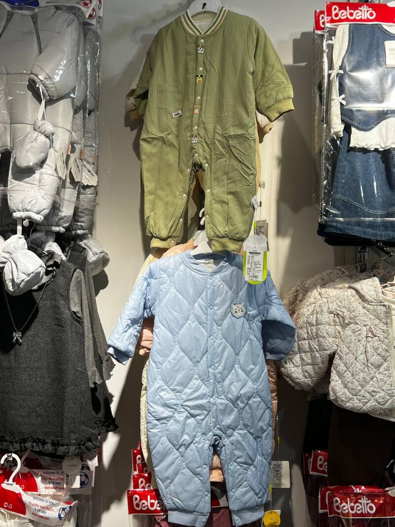 The image shows a display of children's clothing, including a light green jumpsuit with buttons and a light blue quilted jumpsuit, along with other packaged clothing items and the 'Bebetto' brand logo visible on some packaging.