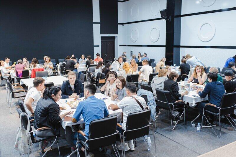A large group of people are sitting at tables covered with white tablecloths, engaged in what appears to be a board game activity in a conference or event setting.