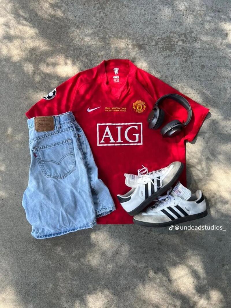 The photo shows a red Manchester United jersey with "AIG" printed on the front, light blue jeans, white Adidas Samba sneakers, and black headphones on a concrete surface.