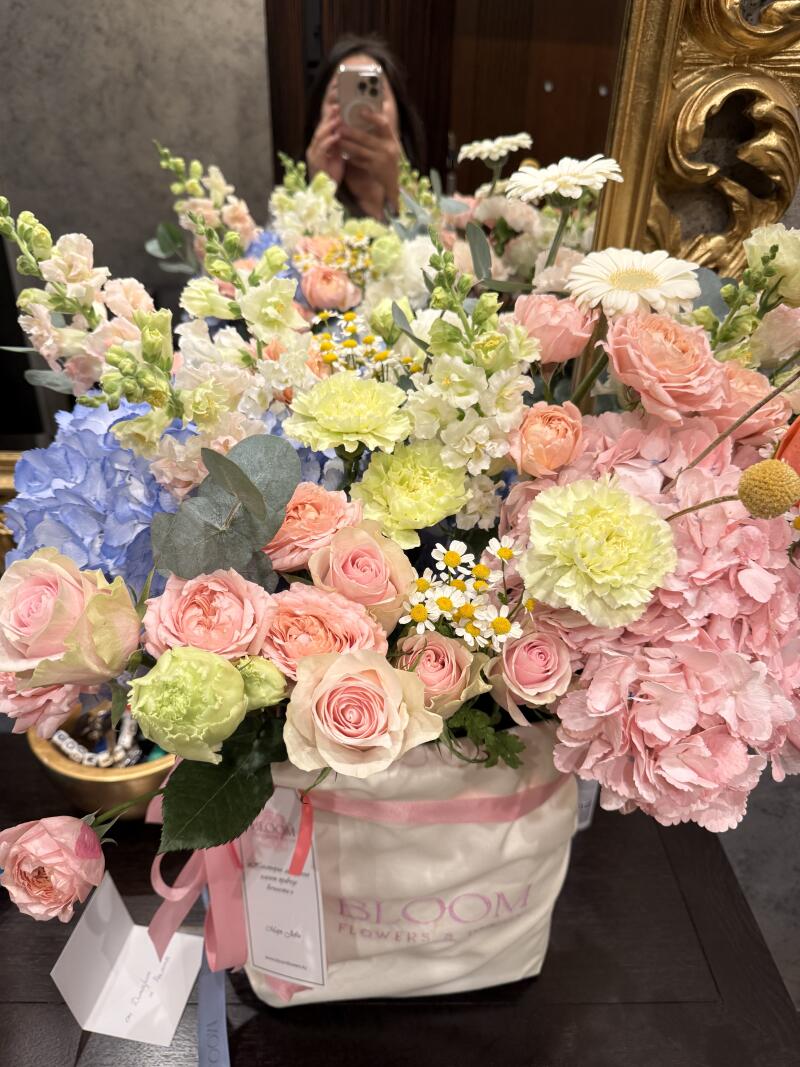 A lush bouquet of assorted flowers, including roses, hydrangeas, and daisies, is arranged in a white "BLOOM FLOWERS" bag with pink ribbon handles, with a person visible in the mirror in the background.