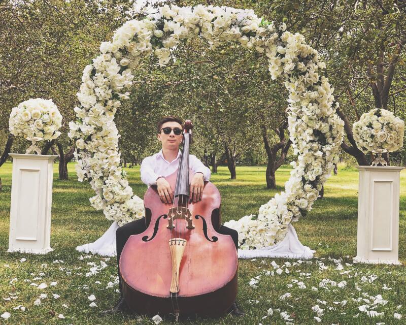 A man in sunglasses sits with a double bass in front of a floral wedding arch in a grassy area with scattered flower petals.