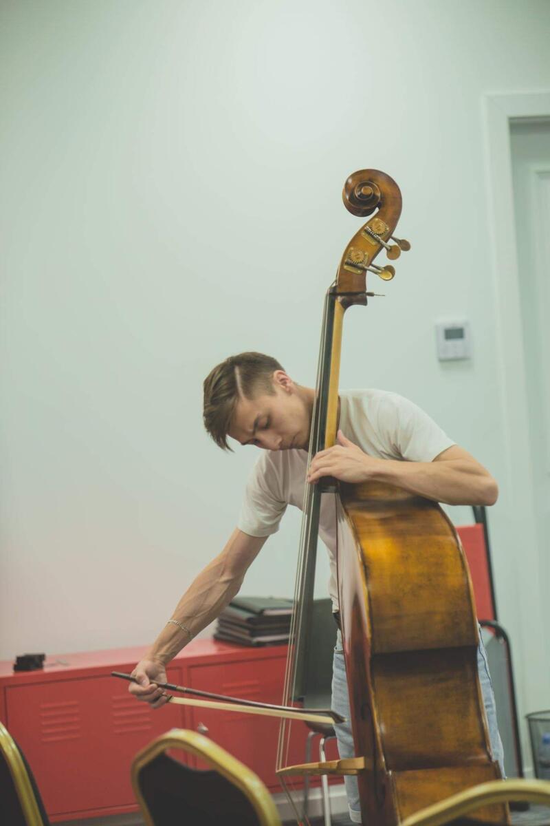 A man in a white shirt is playing a cello with a bow in a room with white walls and a red cabinet.
