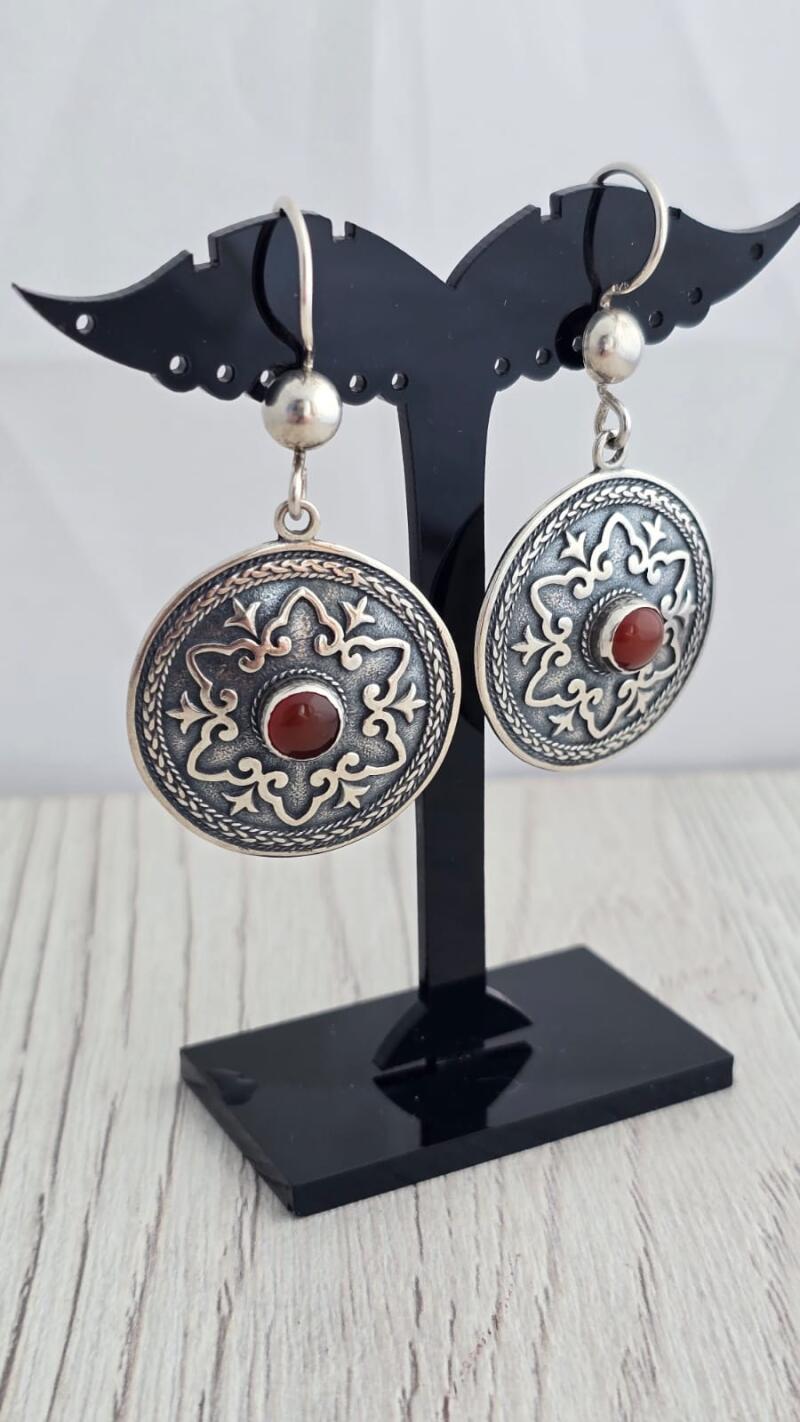 Two silver earrings with red stones are displayed on a black stand, featuring a circular design with ornate patterns.