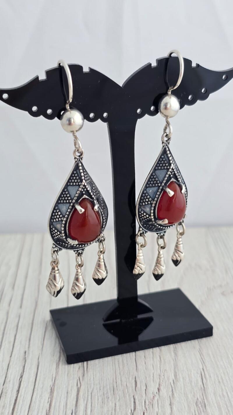 Two teardrop-shaped silver earrings with red stones hang on a black display stand.