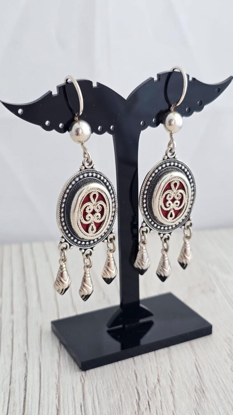 Two silver earrings with red accents hang on a black display stand. Each earring features a circular design with an ornate red center and three small teardrop-shaped pendants.
