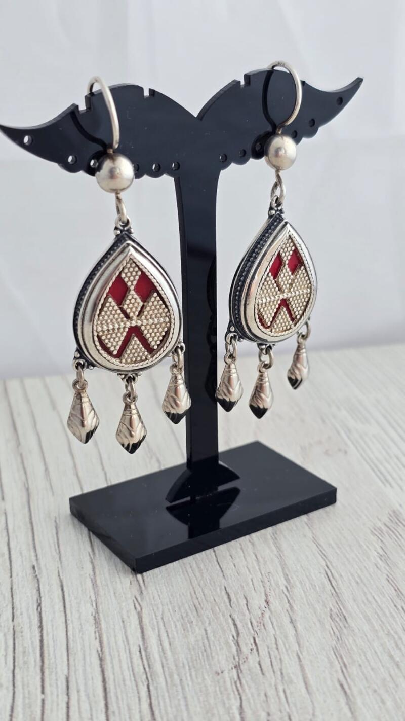 Two teardrop-shaped silver earrings with red and textured silver patterns are displayed on a black stand against a white wood background.